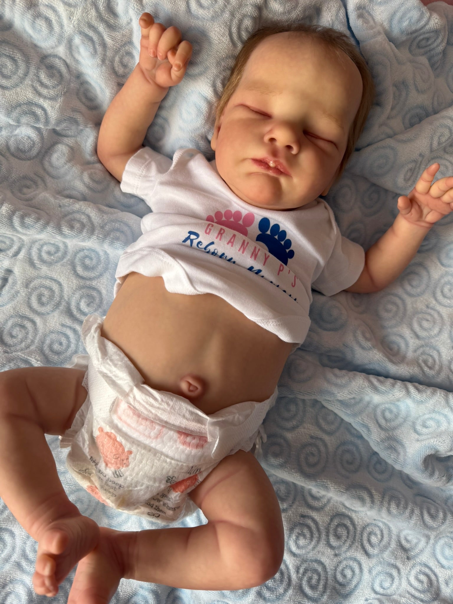 Newborn baby lying on a textured blanket wearing a white onesie with text.