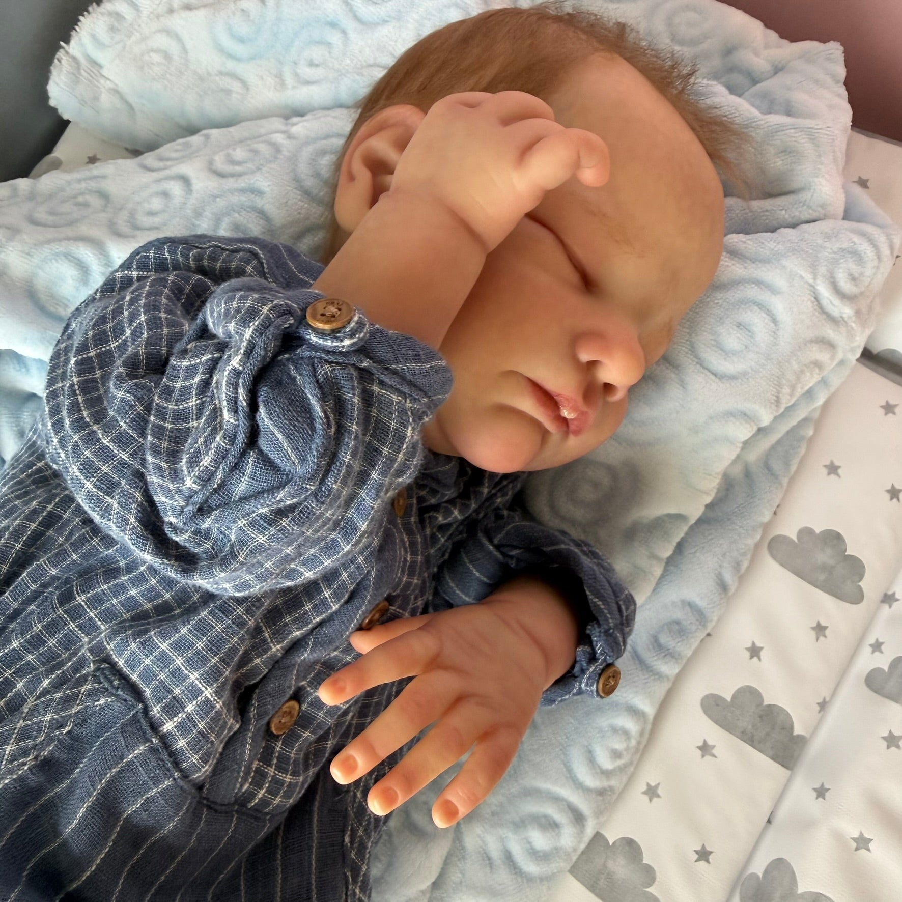 Realistic baby doll wearing a blue checkered outfit on a soft surface with cloud patterns.